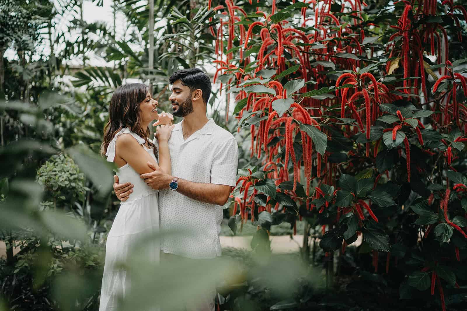 Elegant couple in white attire embracing amidst lush tropical greenery and vibrant red flowers, perfect for a luxury wedding celebration.