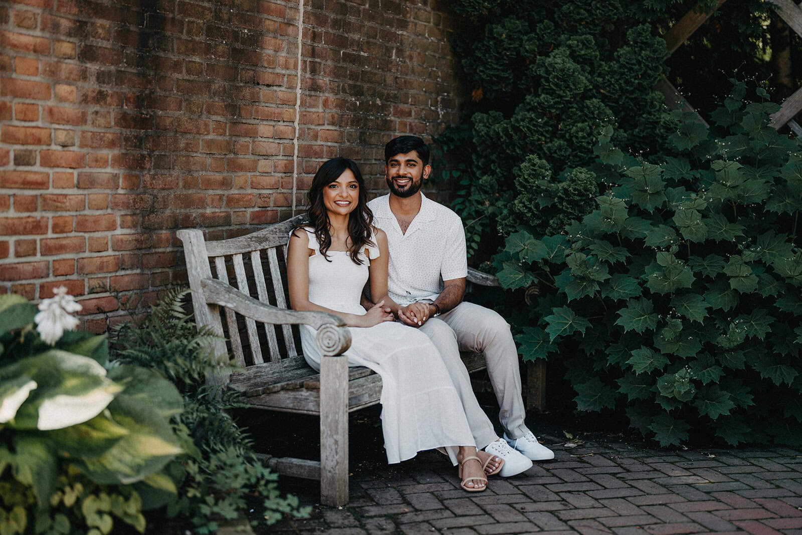 Elegant couple sitting on a rustic wooden bench in a lush garden for a luxury wedding photoshoot.