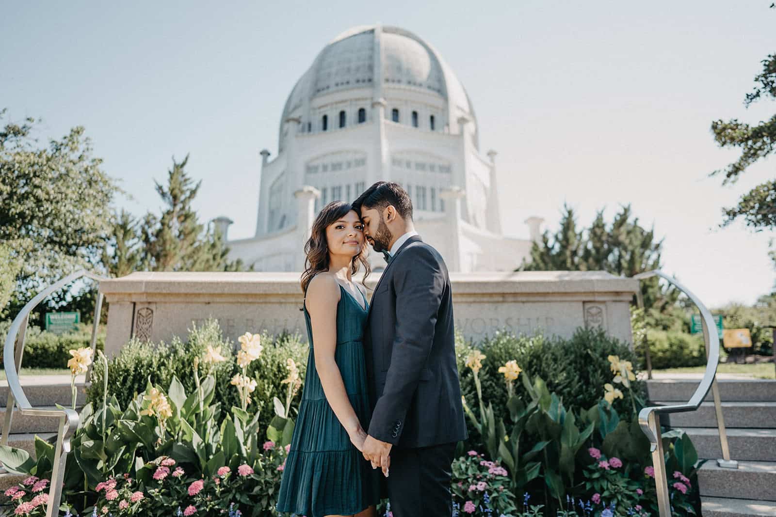 Couple standing close together in front of a grand white mansion, luxury wedding venue, elegant outdoor setting with flowers.