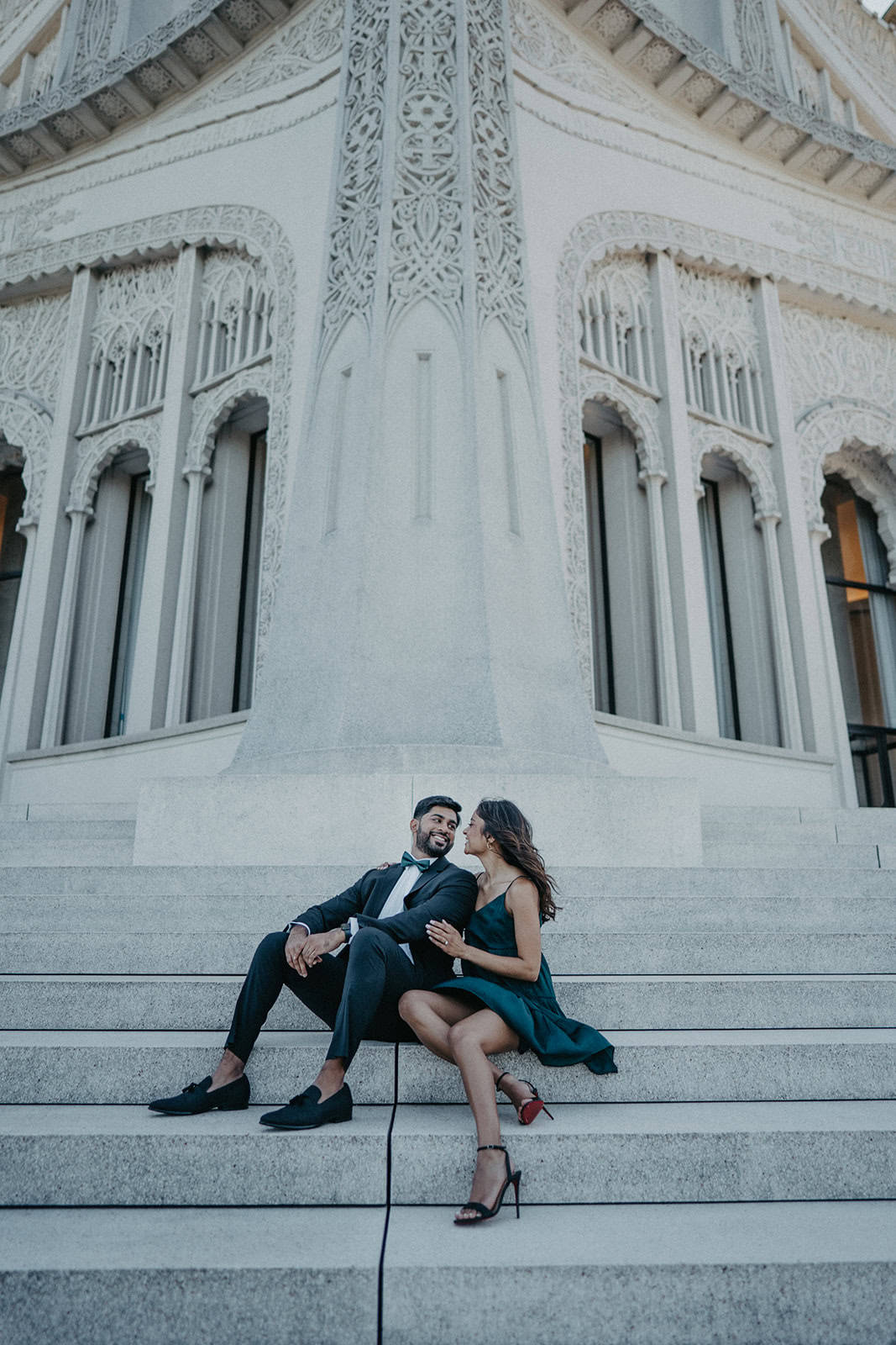 Elegant couple in formal attire sitting on grand stone steps outside a luxurious ornate building, perfect for luxury wedding photography.