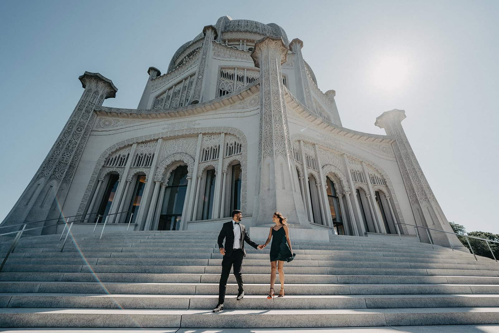Elegant couple holding hands on grand marble staircase in front of white ornate palace, perfect for luxury wedding venues.