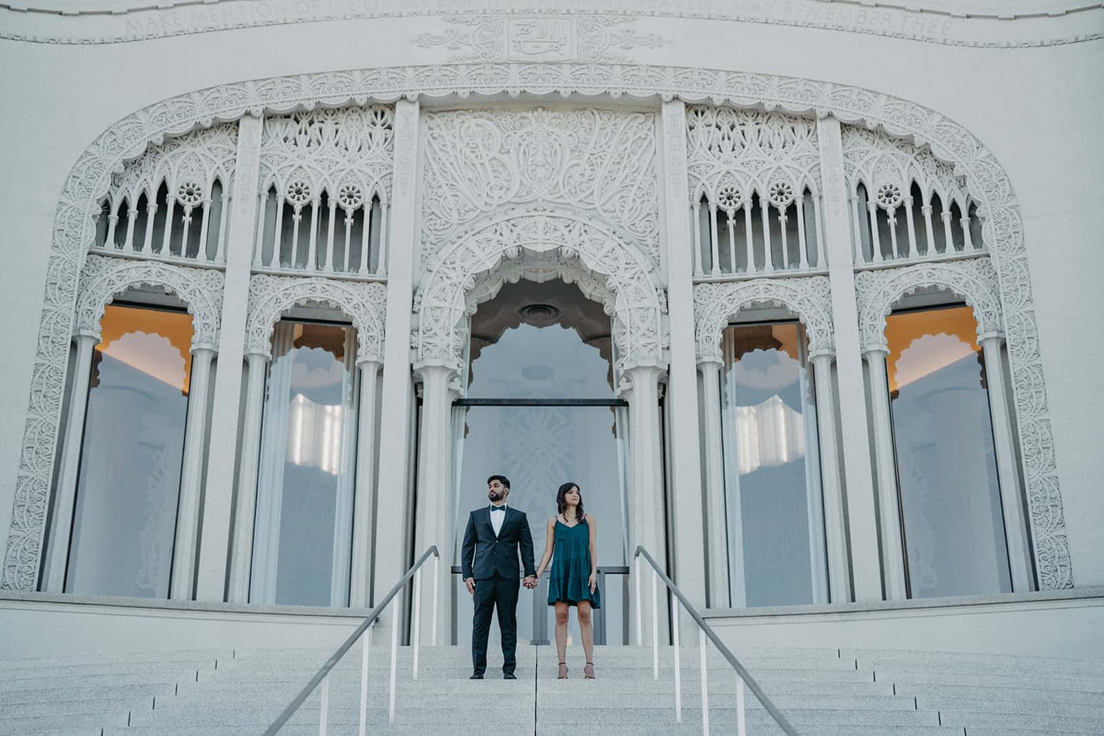 Elegant couple holding hands on grand staircase in front of white luxury wedding venue with detailed architectural carvings.