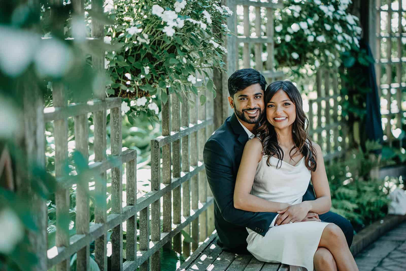 Elegant couple sitting on wooden bench in lush garden setting.