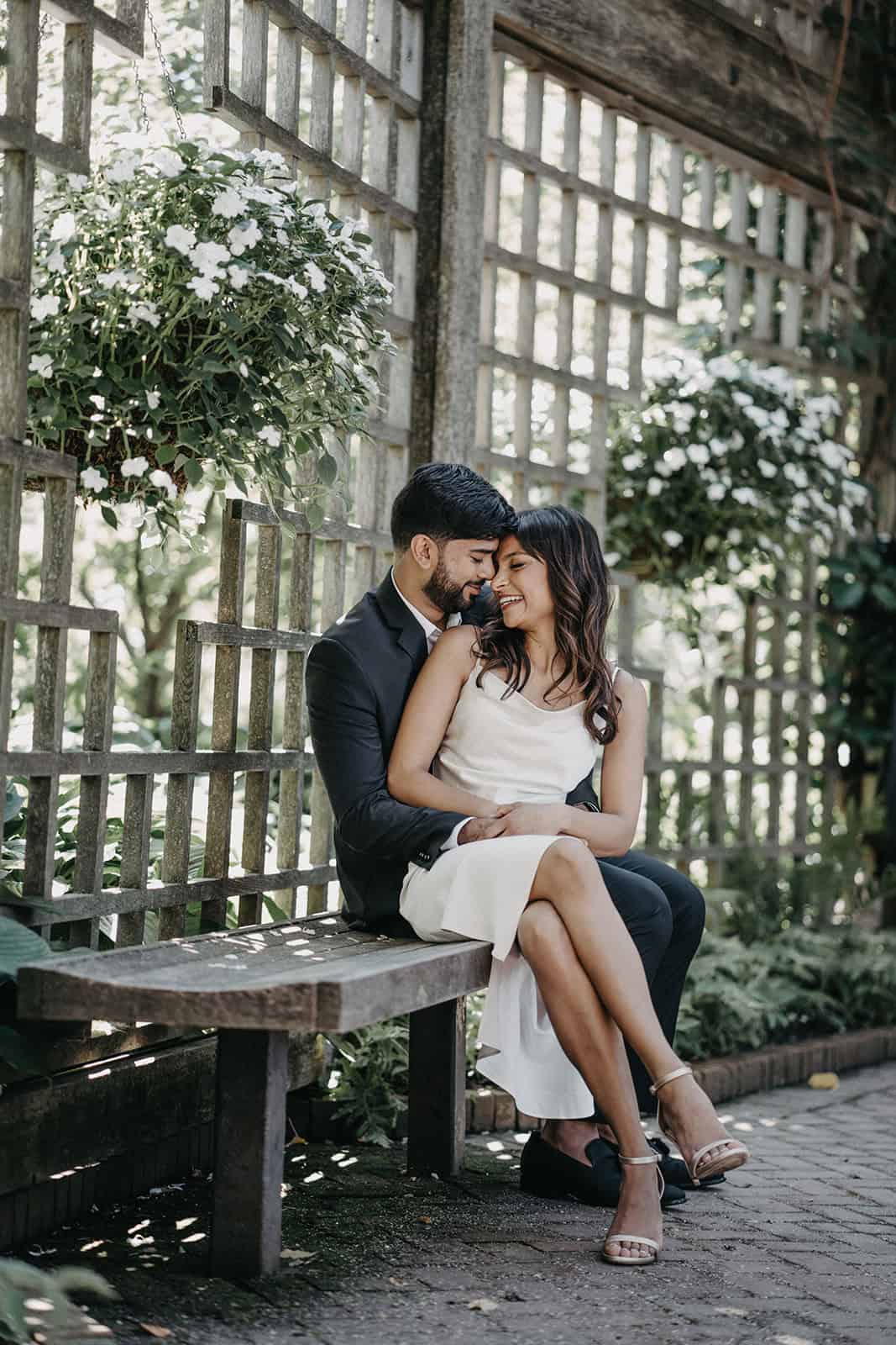 Romantic couple sitting on outdoor wooden bench amidst lush greenery and white flowers, elegant wedding attire, intimate moment, natural setting, luxury wedding venue, high-end outdoor location.