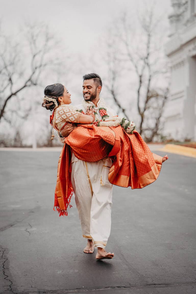 Elegant couple in traditional attire at a luxury outdoor wedding venue, with the groom carrying the bride on a paved street, surrounded by classic architecture and leafless trees.